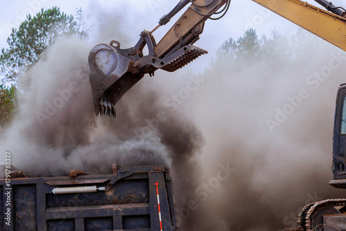 Excavator is lifting dirt debris from ground into dump truck, creating dusty cloud