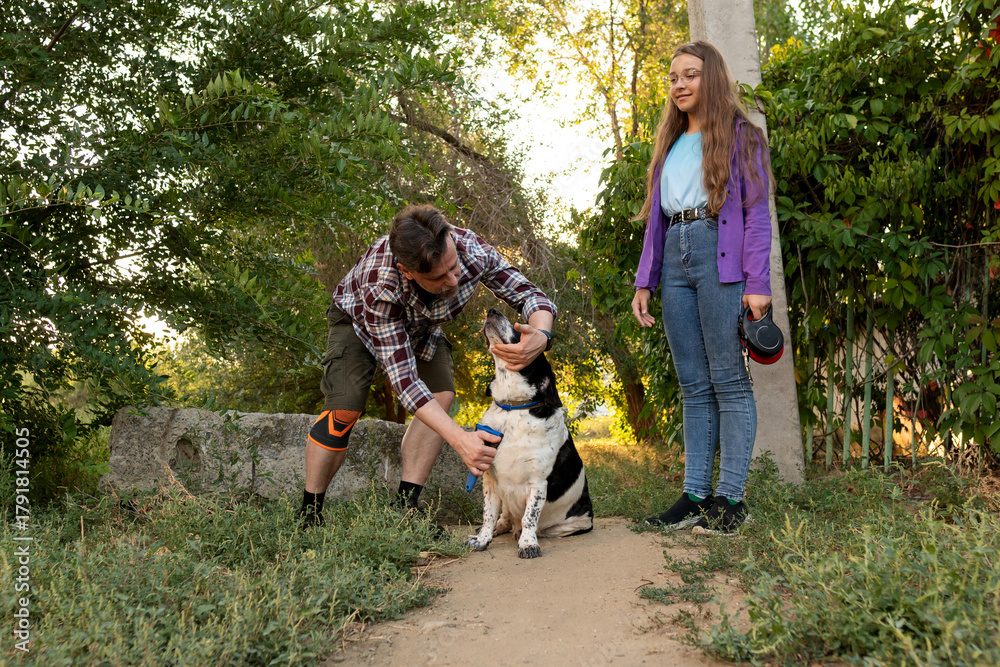 Fototapeta premium Man brushes his black and white dog sitting calmly on a garden path, while a young girl with long hair and glasses stands close, holding the leash, watching with care and affection outdoors