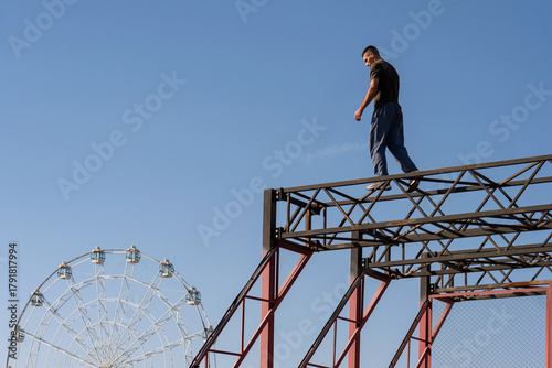 A freerunner walks on a narrow steel beam above the park, with a Ferris wheel in the background. The scene blends urban athleticism with a leisure park environment