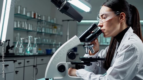 technologist peering through microscope with safety glasses, focused observation of biological slide, glassware and reagent bottles aligned on benches, quiet analytical atmosphere and precise