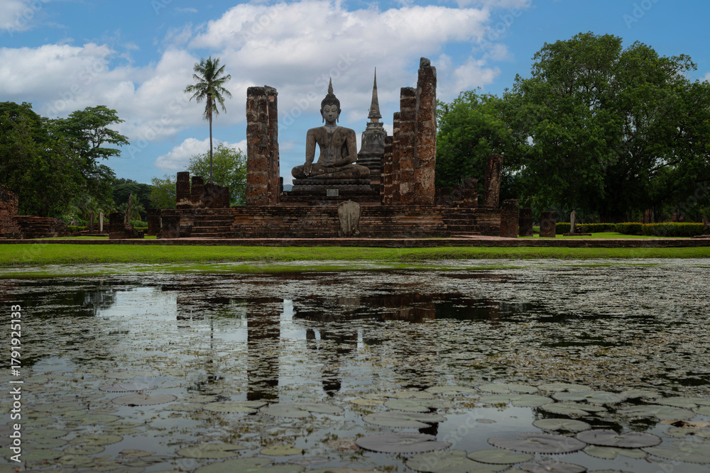 Obraz premium A serene Buddha image stands amid brick ruins beside a lotus-filled pond, creating a tranquil historical sanctuary at Sukhothai Historical Park.