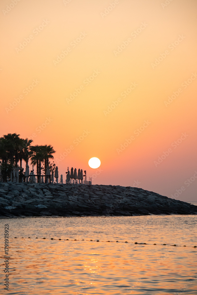Fototapeta premium A large, bright sun setting or rising over a dark, rocky breakwater, silhouetting palm trees and folded beach umbrellas against a vibrant orange and yellow sky