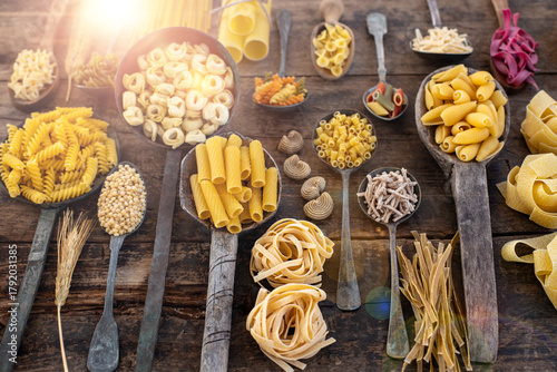 Different types of uncooked pasta on rustic wooden table, in the spoons, cutting board and bowls 