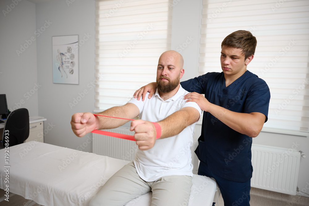 Fototapeta premium Physiotherapist guiding male patient with resistance band exercise for shoulder rehabilitation in clinic. Physical therapy session focused on injury recovery, strengthening, mobility improvement