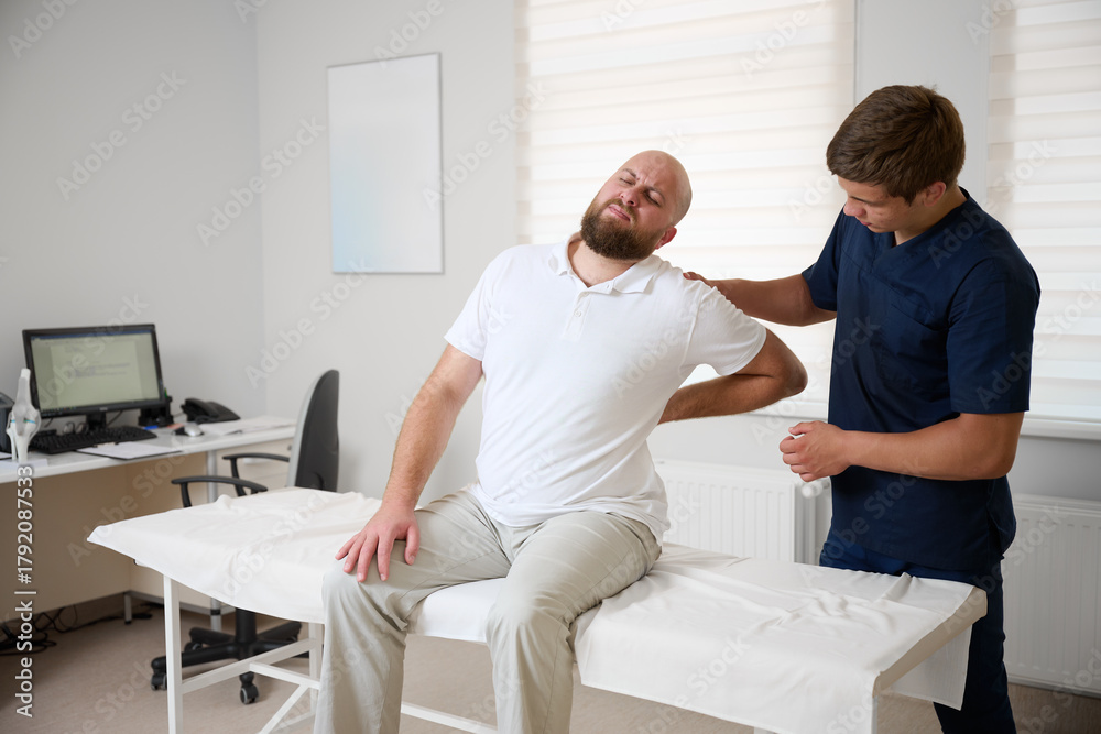 Fototapeta premium Man suffering from back pain sitting on examination table in physiotherapy clinic. Concept of spine problems, rehabilitation, and medical consultation.