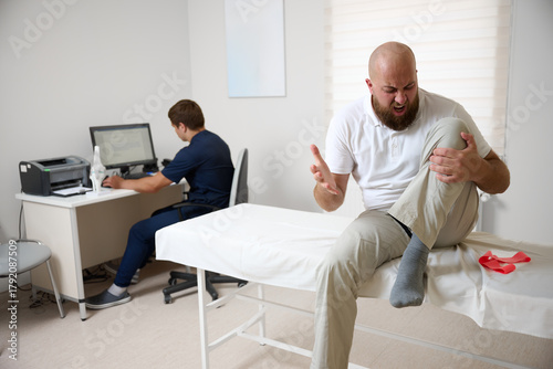 Male patient being examined in clinic for knee pain. Male suffering from knee pain osteoarthritis. Close up man holding his knee due to physical injury while sitting in room.