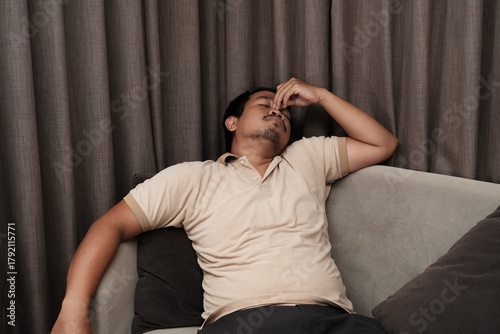 Southeast Asian man in a beige shirt leaning back on a sofa, covering his mouth with one hand while yawning, showing tiredness or boredom.