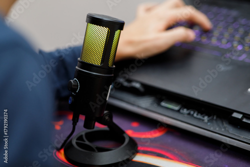 Close-up of a professional usb condenser microphone with yellow light, in use near a laptop keyboard and a hand typing