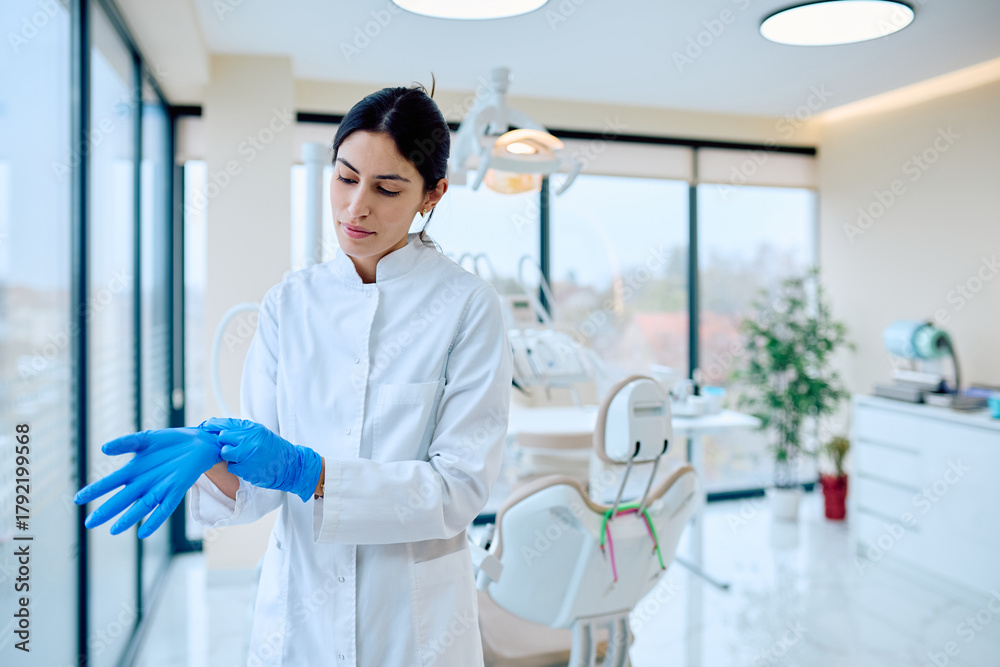 Fototapeta premium Female dentist or assistant preparing for work, putting on blue protective gloves in a modern dental clinic or office