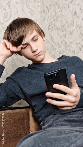 Disappointed sad teenage boy sitting on sofa using smartphone, playing video game. Scrolling social media, surfing internet at home alone. Sad child on coach in living room with phone.
