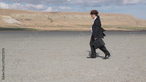 A curly-haired teenager with glasses and a mature business suit stubbornly walks forward, shielding himself from the wind with his hand, against the backdrop of a hill and blue sky
