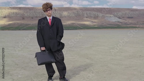A sad teenager in a mature business suit with a briefcase stands on dry ground against the backdrop of a hill with a vineyard. The camera zooms out