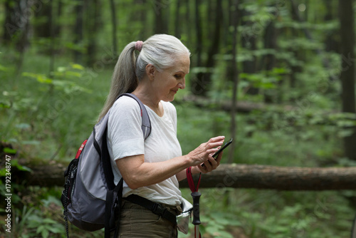 senior woman  with  a backpack uses a mobile phone standing on in the summer forest. copy space. mental health. Slow life.  