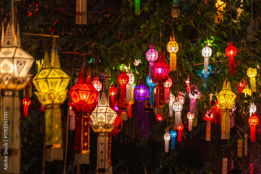 Naklejka premium Vibrant traditional Lanna lanterns illuminate the night, hanging amidst lush foliage. This festive display captures the enchanting atmosphere of a cultural celebration, possibly during Thailand's Yi