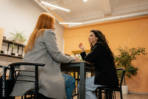 Two professionals share a casual conversation across a table in a bright, contemporary workspace.