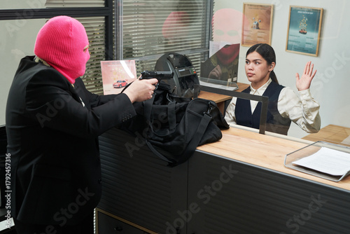 Caucasian man wearing pink ski mask pointing handgun at young adult Hispanic woman behind counter raising hand during bank robbery, office posters and documents visible in background