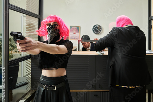 Young adult Caucasian woman with pink wig and bandana holding handgun and aiming forward during bank robbery while young adult Caucasian man in pink mask threatening bank teller in background