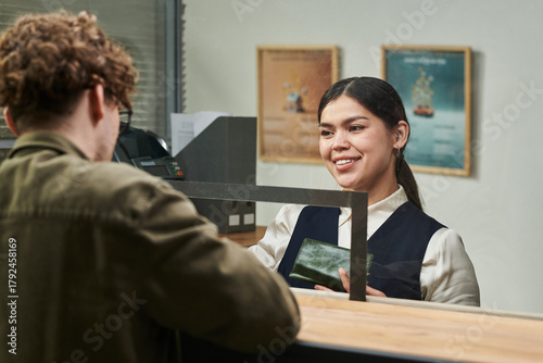 Young adult Hispanic woman smiling while assisting young adult Caucasian man at service counter, holding passport and engaging in customer service interaction through glass partition