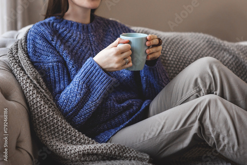Woman sitting and relaxing on sofa at home holding blue mug in hands. Cozy autumn mood with knitted blanket and warm sweater, peaceful moment indoors