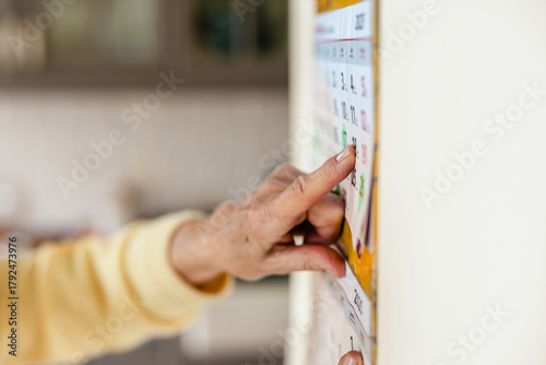 Senior woman using a wall calendar in her kitchen, focus on finger
