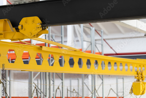 Yellow overhead crane beam with a steel trolley connection to a main I-beam girder in an industrial warehouse. The electric chain hoist and power cables are visible below. Industrial factory interior.