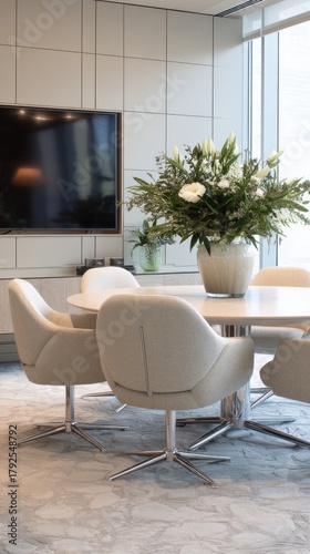 A sleek, modern conference room featuring a round table, comfortable chairs, and a large flower arrangement, with a wall-mounted TV.