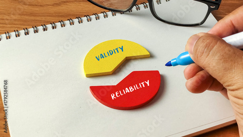 Hand writing next to Validity and Reliability wooden puzzle pieces on a notebook with glasses for research and quality concepts.