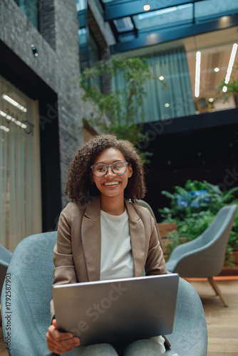diverse female freelancer smiling with laptop in cozy atrium armchair, relaxed posture, remote work setup,