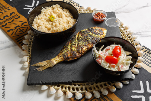 Attiéké servi avec poisson braisé, semoule de pulpe de manioc fermentée cuite à la vapeur, présenté sur une ardoise et un set de table tissé africain – plat traditionnel africain.