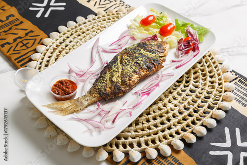 Bild auf Leinwand Poisson braisé, présenté sur une ardoise et un set de table tissé africain – plat traditionnel africain