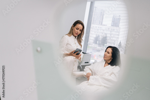 Reflection in mirror of aesthetician doctor consulting middle-aged female client in beauty clinic, showing information on mobile phone for skincare treatment planning in modern medical spa office.