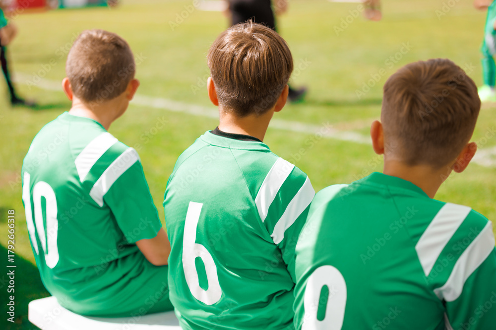 Fototapeta premium Boys in Green Jerseys Sitting on Sideline Bench. Boys Soccer Team Sitting on Bench. Young Players in Green Jerseys Watching Football Game during Youth Sports Event