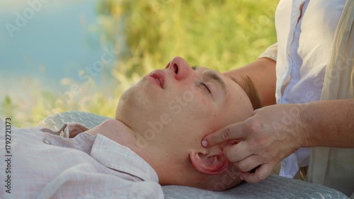 Woman massaging ear and temple of man near river for relaxation. Professional massaging head with calming energy on fresh air. Practitioner working on ear for peace and relaxation by water. Therapist
