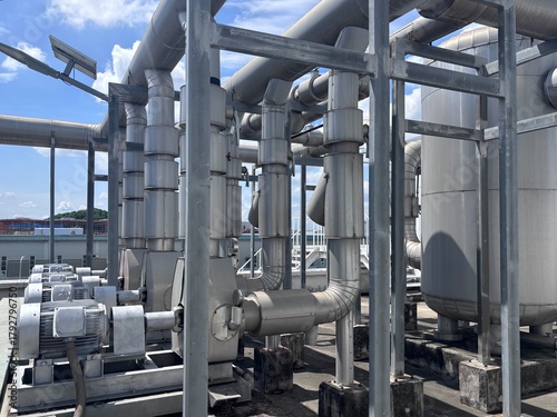 A complex view of silver industrial piping, large valves, and electric motors forming part of a chiller plant or HVAC system on a commercial rooftop. The structure shows essential mechanical infrastru