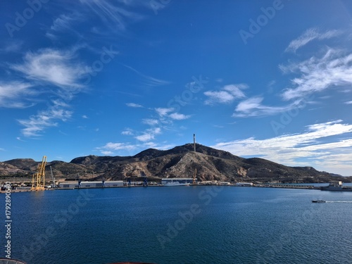 spain marcia jan 4 2024 Harbor Panorama: Hills, Cranes, and Tall Monument Rising Over Calm Blue Sea Under Clear Sky
