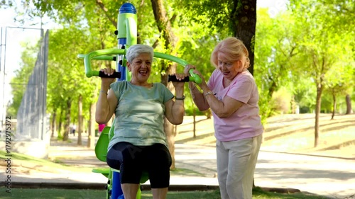 Two cheerful senior women enjoying an active lifestyle, exercising on outdoor gym equipment in a sunny park. One woman uses a shoulder press machine while her friend provides motivation and support
