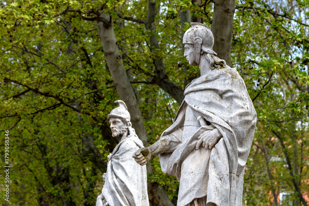 Obraz premium A close-up of two white stone statues of Spanish monarchs wearing classical armor in the gardens of the Palacio Real, framed by green trees and foliage under a bright sky