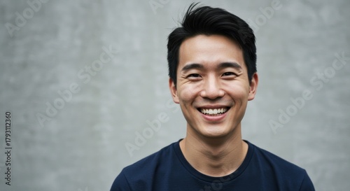 A smiling young Asian man in a dark blue tshirt stands against a textured gray wall