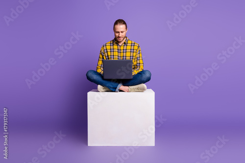 Slika na platnu Young handsome guy sits cross legged on a white block with a laptop in a purple