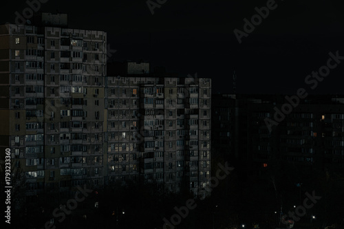 Tall apartment buildings stand in darkness at night, with only a few illuminated windows suggesting occupancy. The scene conveys a somber, possibly blackout-affected urban atmosphere
