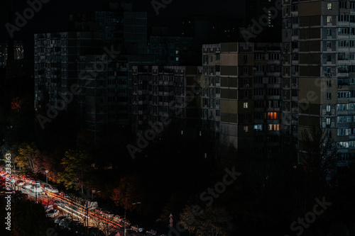 A darkened urban cityscape at night with minimal lights visible in apartment buildings and street traffic. The scene evokes a sense of blackout or power outage