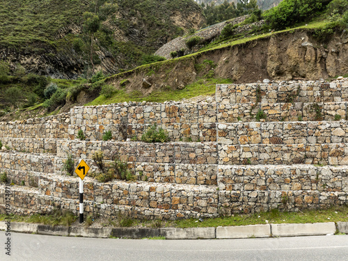 A strong gabion retaining wall, made of stones in wire baskets, provides stability and prevents erosion on a mountain roadside next to a yellow curve sign.