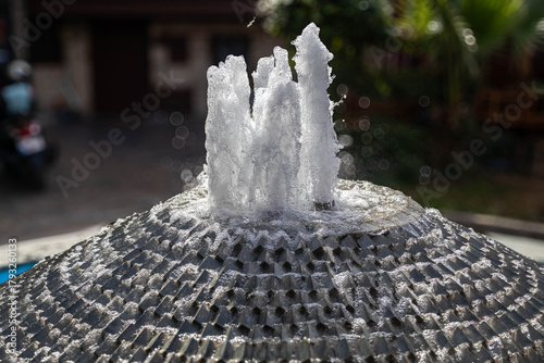 Fototapeta Naklejka Na Ścianę i Meble -  Backlit water splashes rise from a small outdoor fountain, glowing in strong sunlight. Clear drops, textured surface and soft bokeh create a vivid scene. Antalya, Turkey.