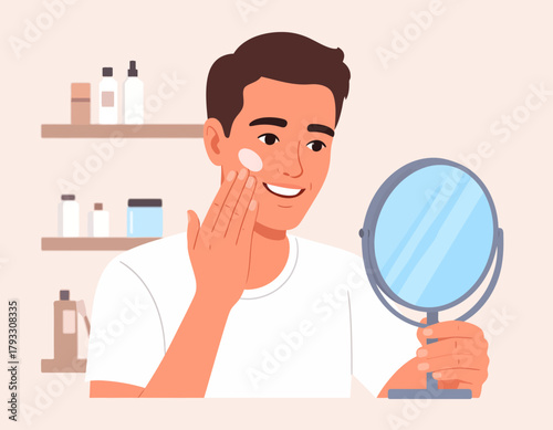 Young man applying face cream while looking in a handheld mirror, with toiletries on a shelf in the background.