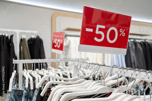 Bright red sale signs indicating a 50 percent discount are prominently displayed in a clothing store. Rows of hangers with various garments create an inviting shopping atmosphere.