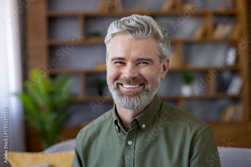 Smiling mature man with grey hair and beard, sitting at home in smart-casual shirt, confident and relaxed for a friendly portrait headshot with warm, natural expression