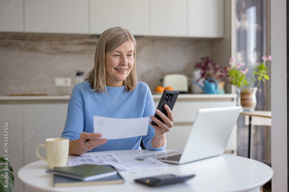 Fototapeta premium Senior woman is managing her personal finances and paying bills online using a smartphone and laptop at home, smiling while organizing household expenses and financial records