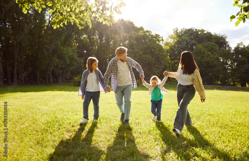 Fototapeta premium Four family members having fun at park during sunny summer day, walking together and holding hands, having good relationships, enjoying nice day. Parents and children resting outdoors