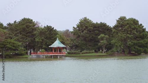 Slow motion showing a row of lakeside trees and red pavilion reflected in smooth lake surface at Ohori Park, Fukuoka, Japan, under soft daylight and natural serenity
