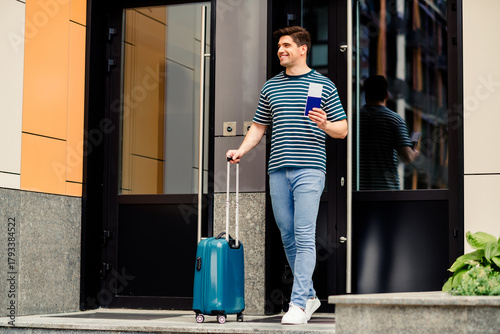 Young man exiting modern building carrying suitcase and wearing striped casual outfit on a sunny day
